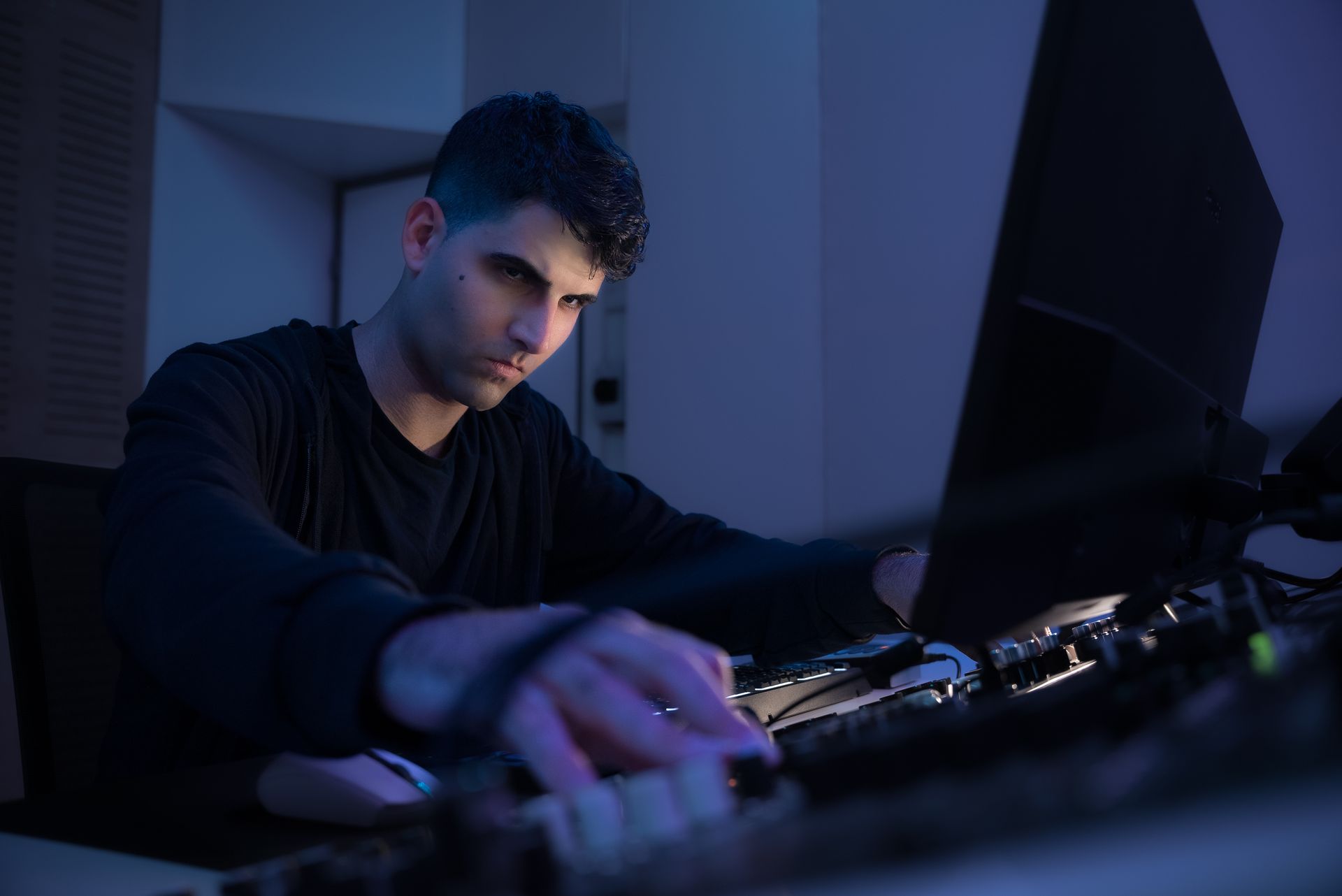 Man in dark room, illuminated by blue light, working on music production equipment.