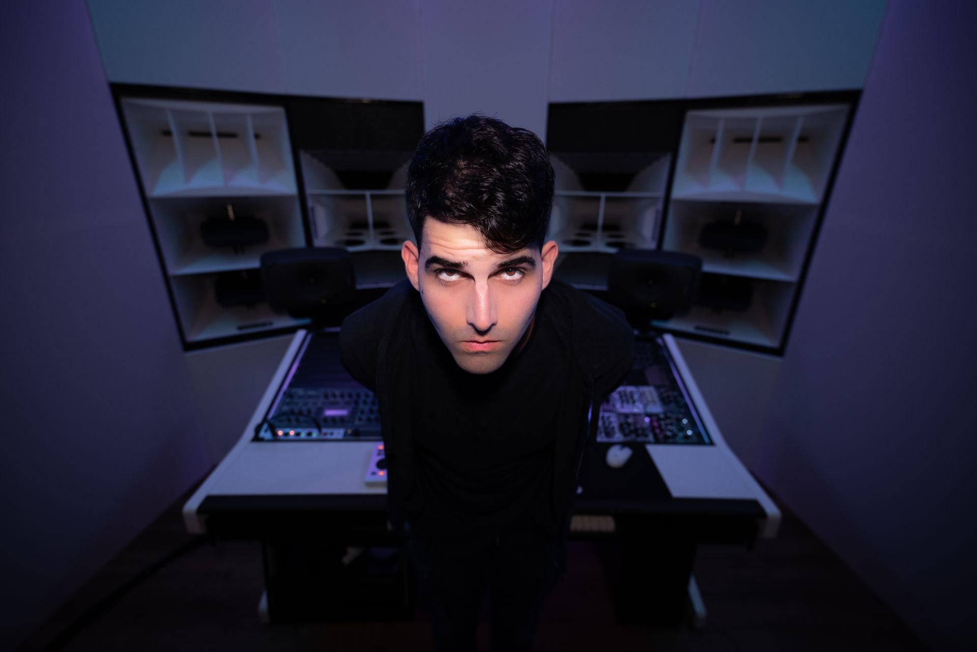 Mann im schwarzen Hemd starrt in einem Musikstudio. Er steht vor einem Mischpult mit großen Lautsprechern.