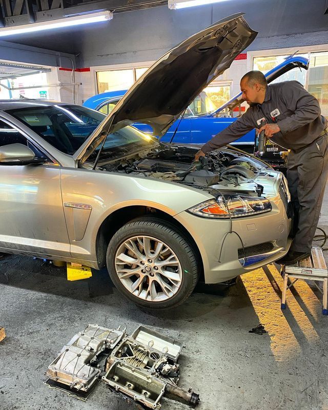 Mechanic working on a silver Jaguar with the hood open, engine parts on the floor, in a garage.