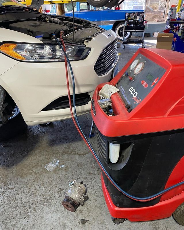 A white car being serviced at a repair shop, connected to a red AC recharge machine. An old part sits on the ground.