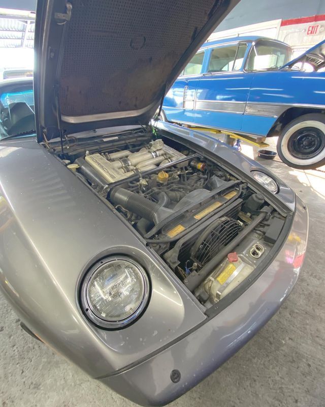 Gray Porsche 928 with hood open, showing the engine. A blue classic car is visible in the background.