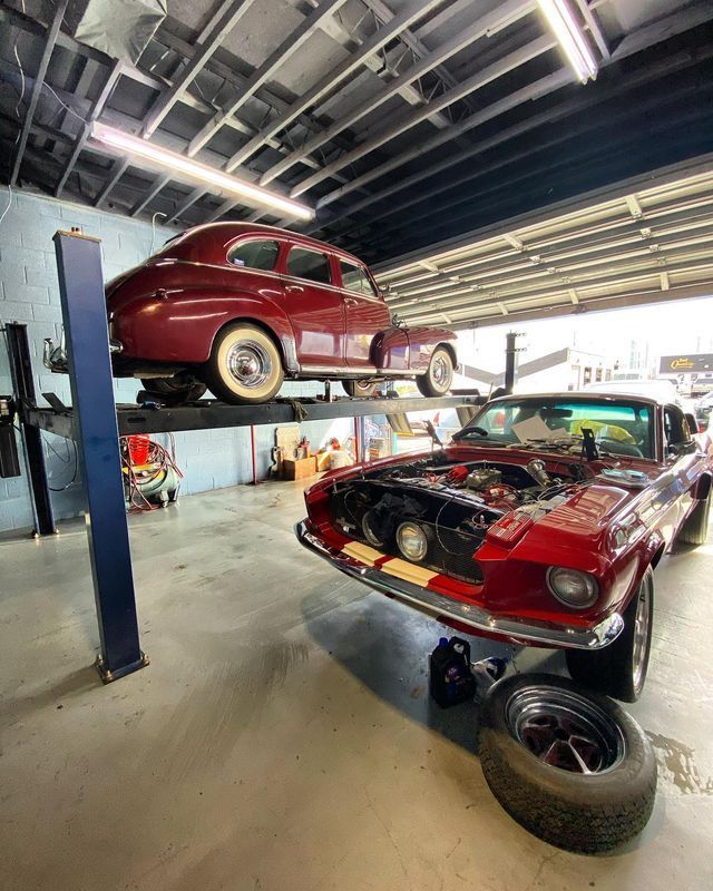 A classic maroon car on a lift over a red car with its hood open in a garage.