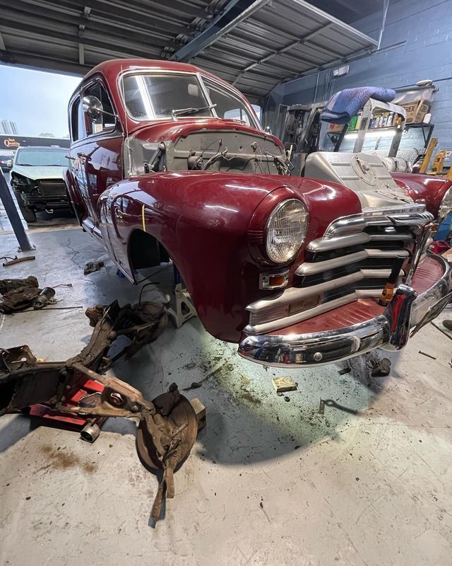 Maroon vintage car undergoing restoration in a garage; front end lifted.