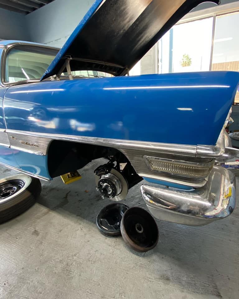 Blue classic car with hood open, wheel components visible, in a repair shop.