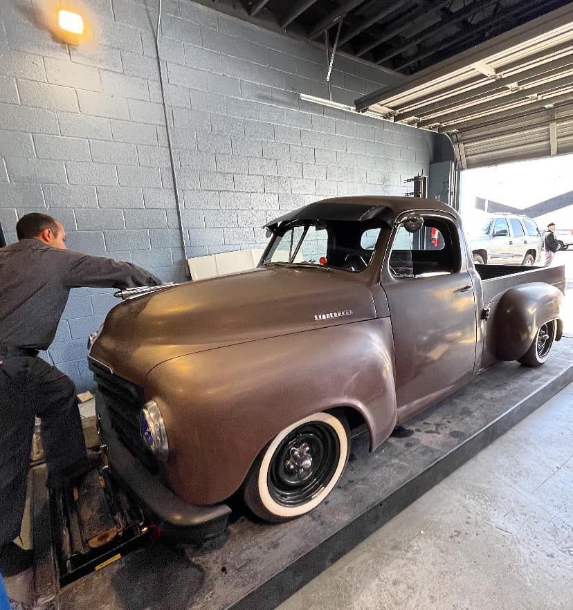 Brown vintage pickup truck at a mechanic shop, being worked on. Mechanic is leaning on the truck.