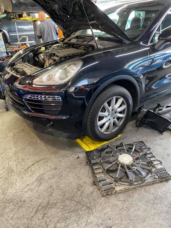 Dark blue Porsche SUV with open hood, being worked on in a garage. Fan on the floor.