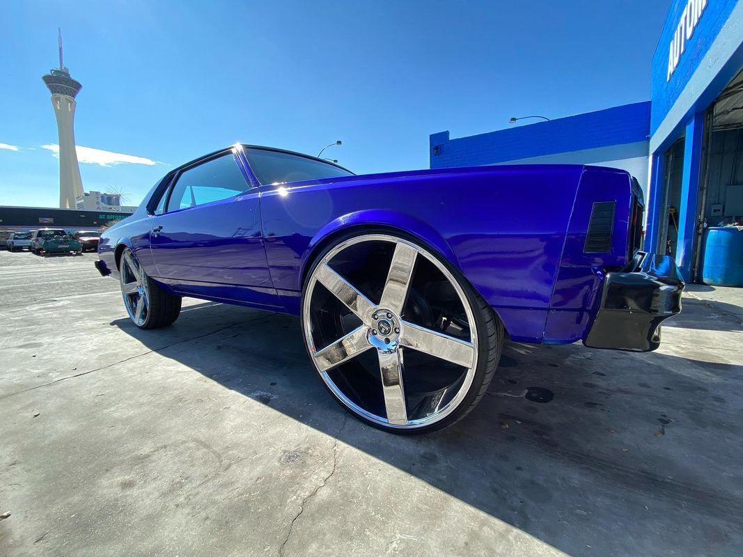 Purple car with large chrome wheels parked outside on a sunny day. The Stratosphere tower is visible in the background.