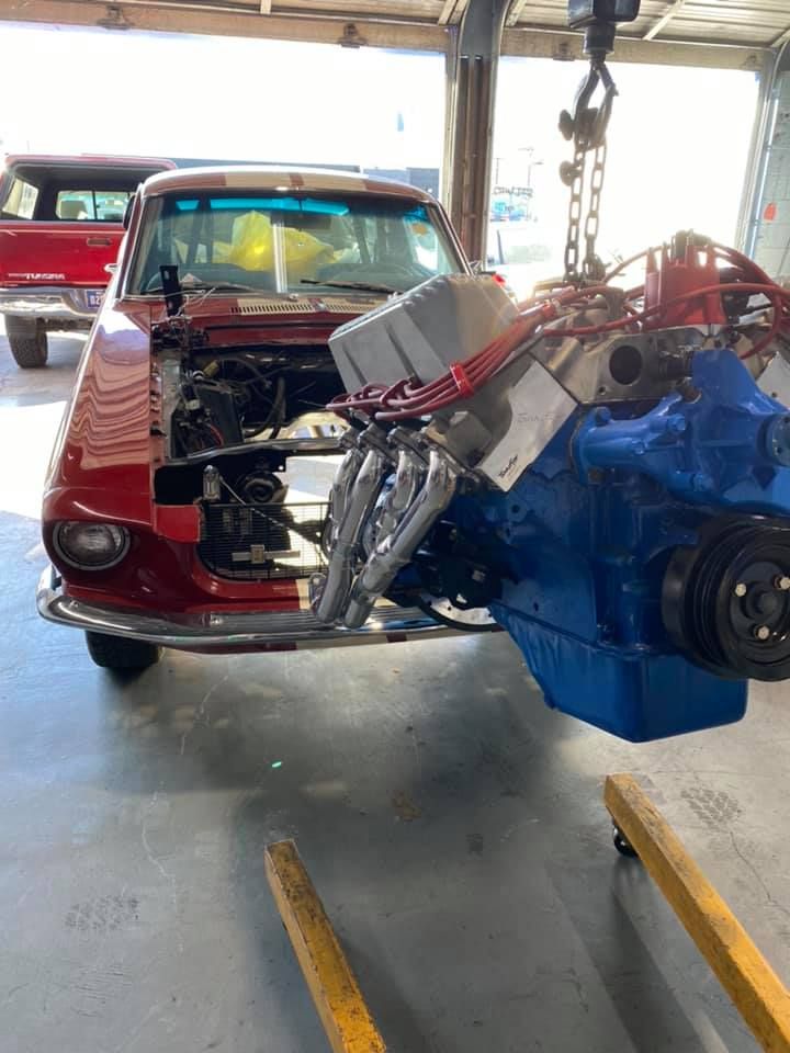 A red classic car with its engine being lowered into the engine bay. A blue engine is suspended by a chain.