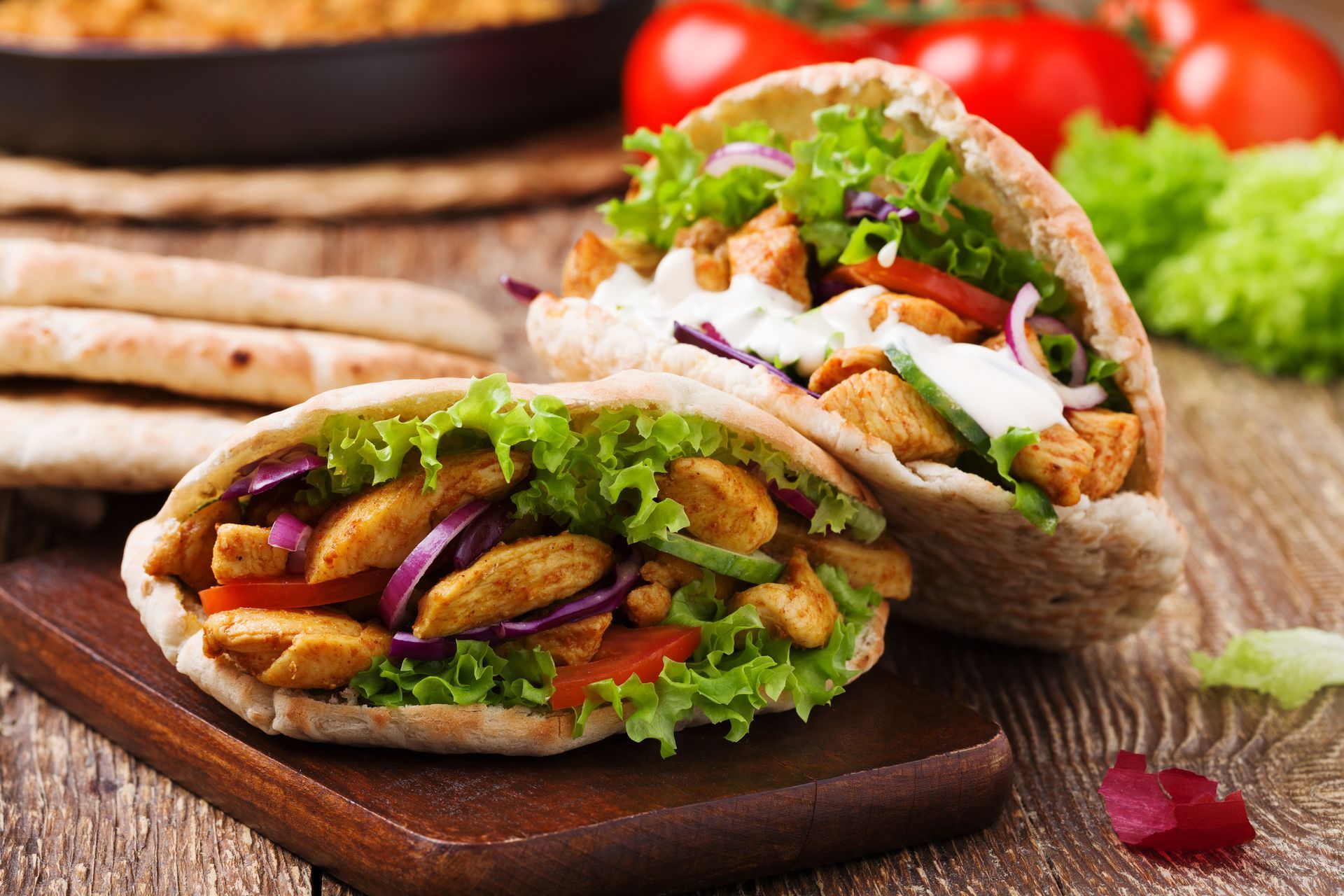 Two pita bread sandwiches with chicken , lettuce , tomatoes and onions on a wooden table.