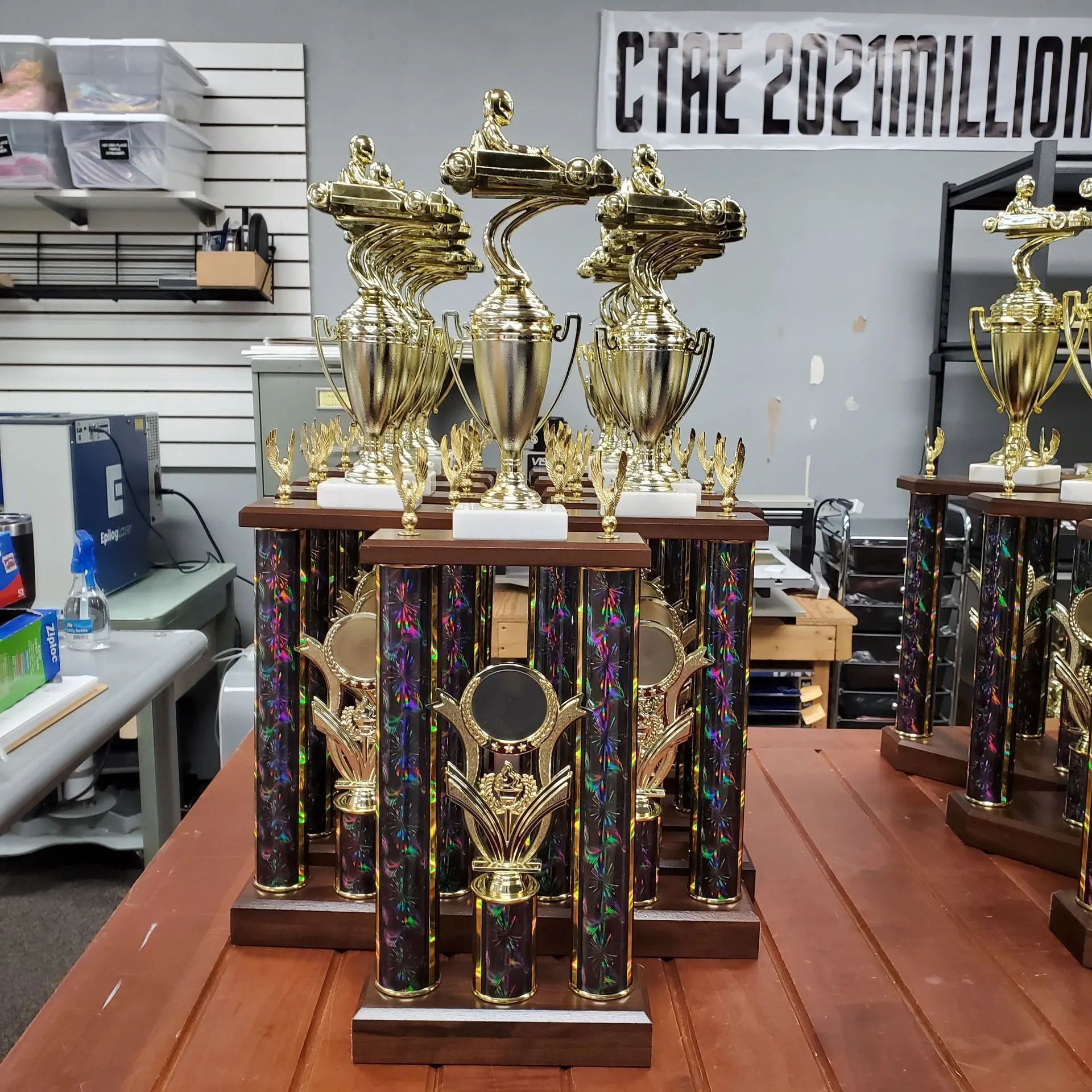 Trophies on a wooden table, with gold cups and detailed bases, in a room with shelves and equipment.