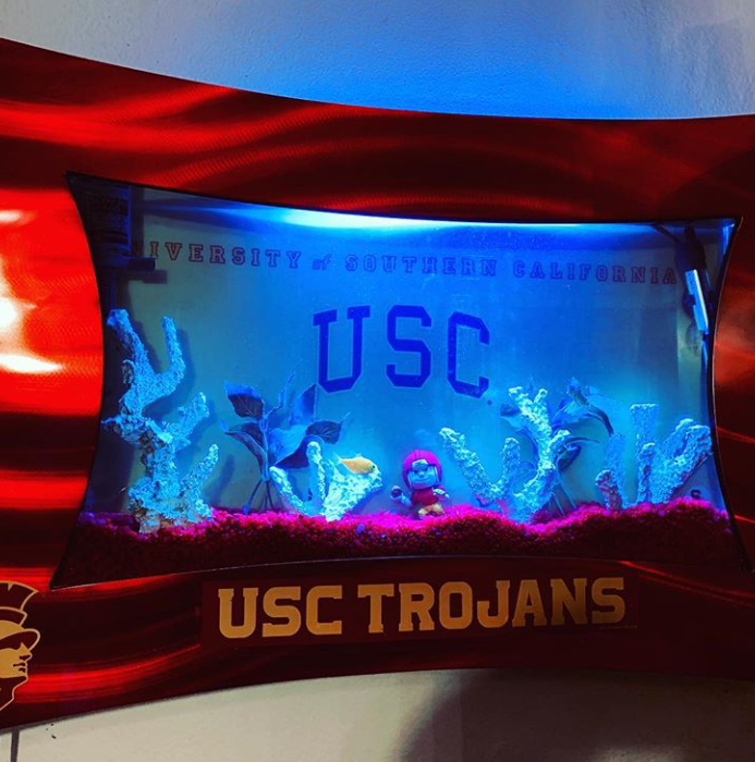 USC Trojans-themed fish tank with coral, decor, and a mascot figure, lit with blue light against red.