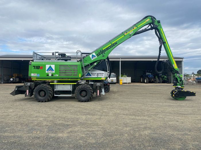 A Green Excavator is Parked in a Gravel lot in front of a Building — Tree Services In Kelso, NSW