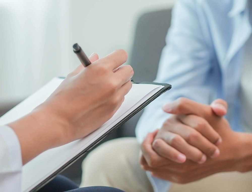 Eine Person schreibt in einem Patientengespräch auf einem Klemmbrett und steht einer anderen Person mit gefalteten Händen gegenüber.