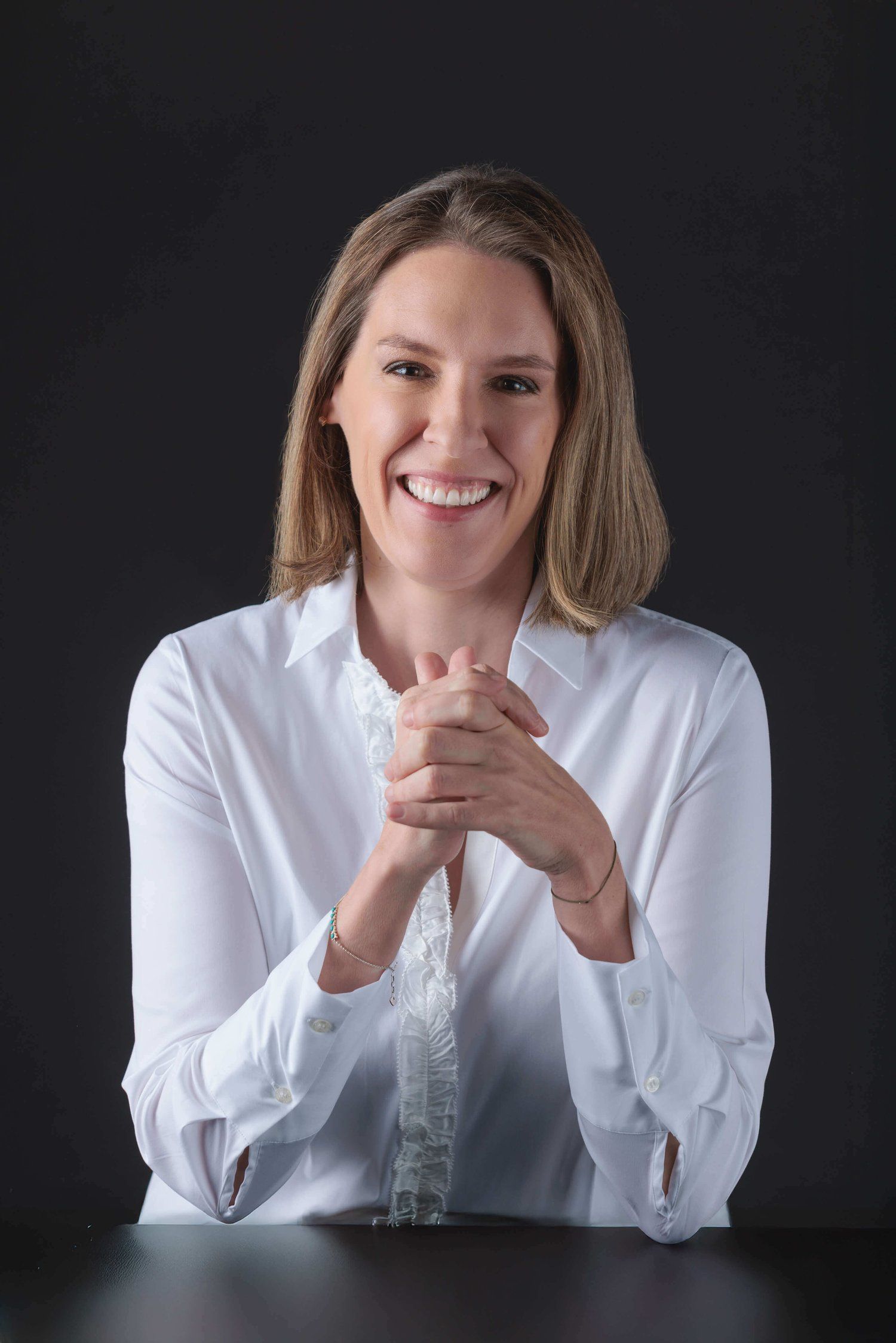 Eine Frau sitzt mit gefalteten Händen an einem Tisch und lächelt.