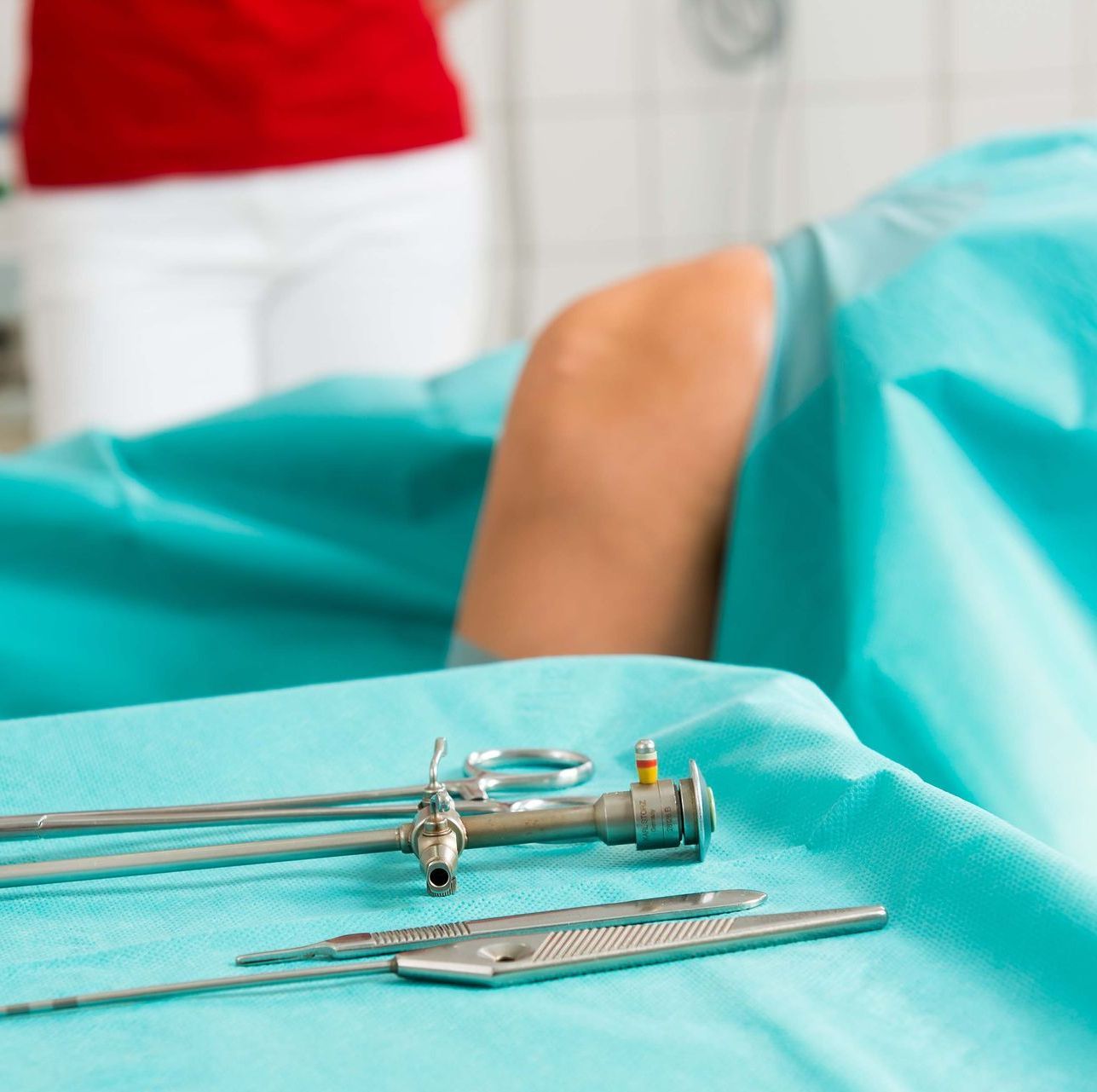 Chirurgische Instrumente auf einem blaugrünen Tisch, im Hintergrund das Bein eines Patienten und ein Arzt in einem roten Hemd.