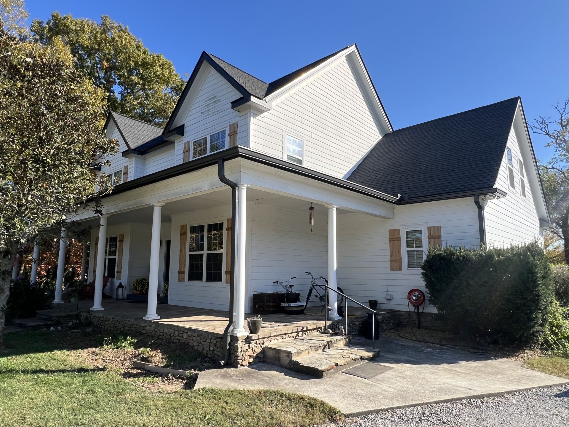 White two-story house with a porch and black roof. Sunny day, surrounded by trees and grass.