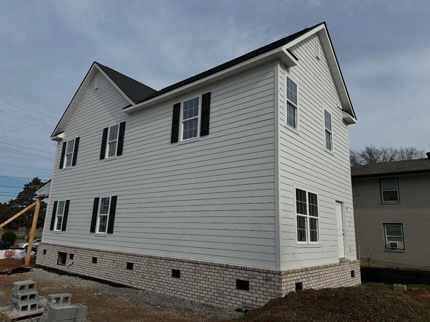 Two-story white house under construction with black shutters and brick foundation; cloudy sky.