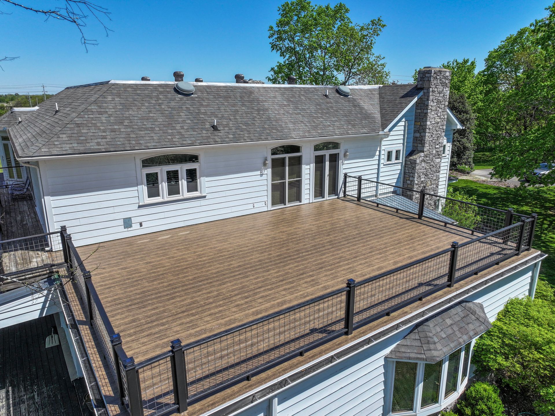 Exterior view of a white house with a large brown deck and black railings. Chimney and trees visible.