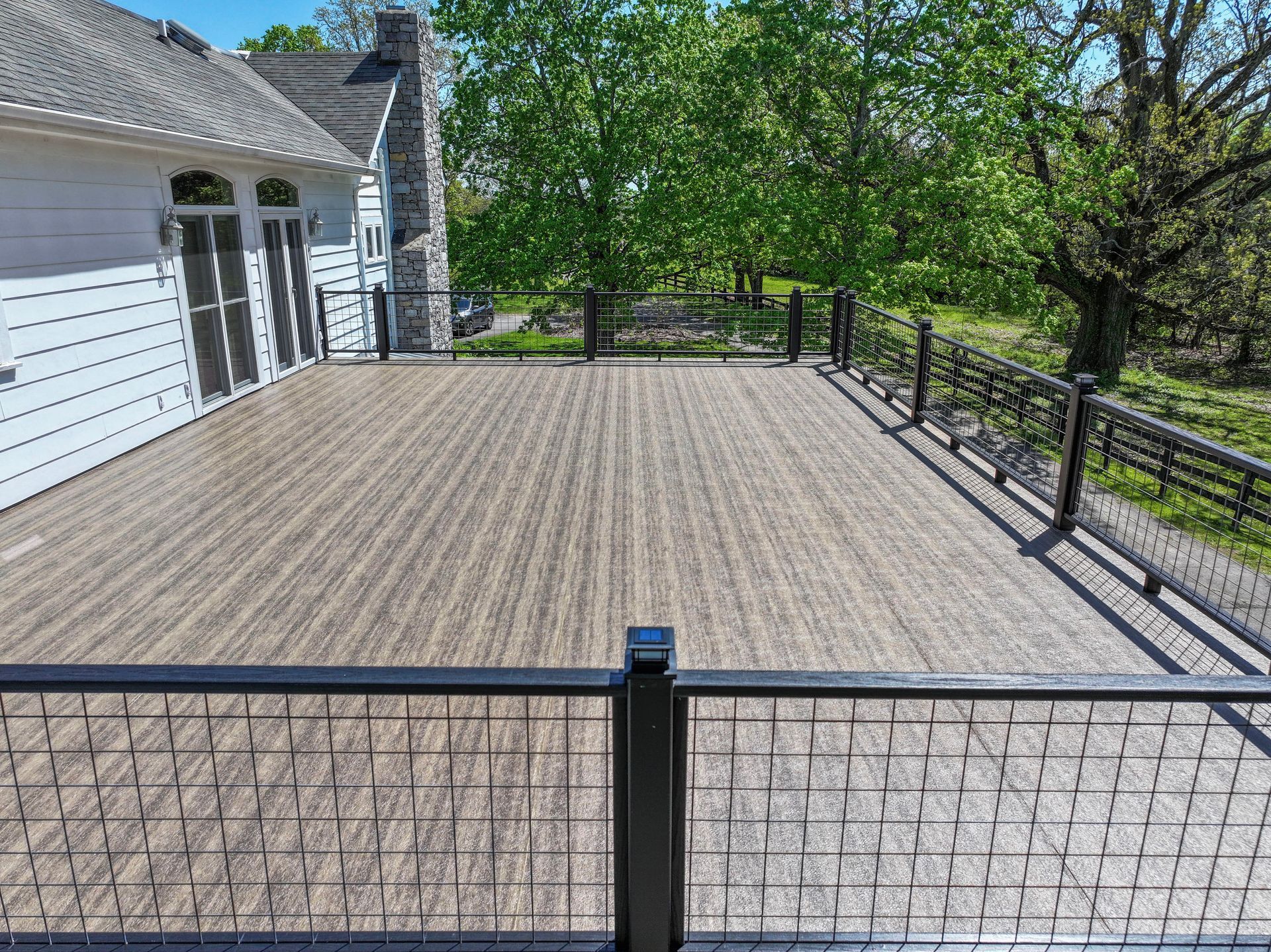 Large wooden deck with black railings overlooking a grassy area and trees under a sunny sky.