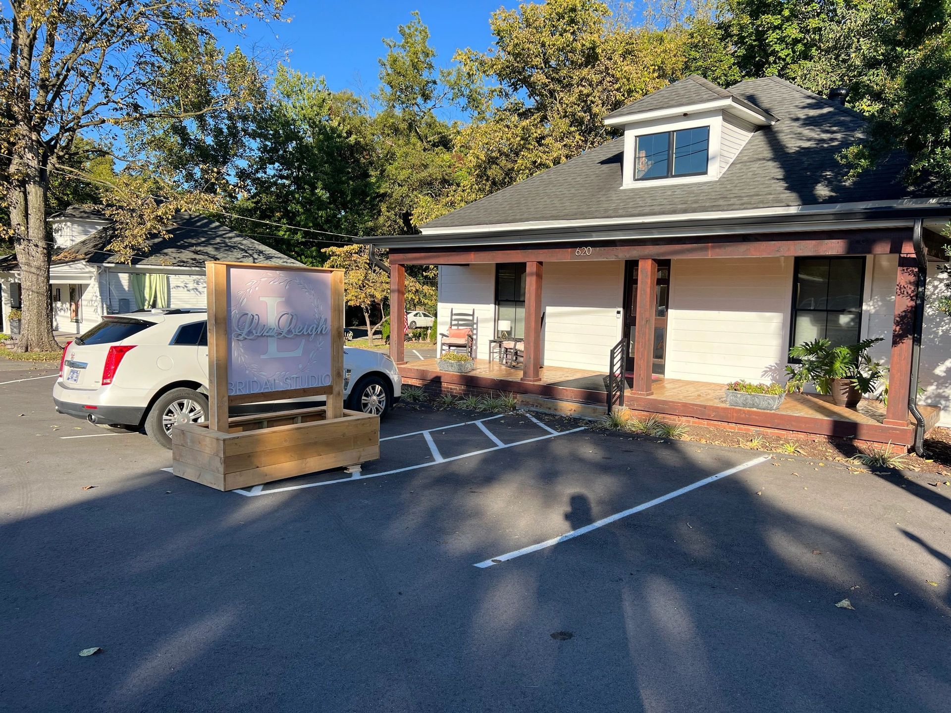 Small white building with porch and sign in front, parked car to the left, trees in background.