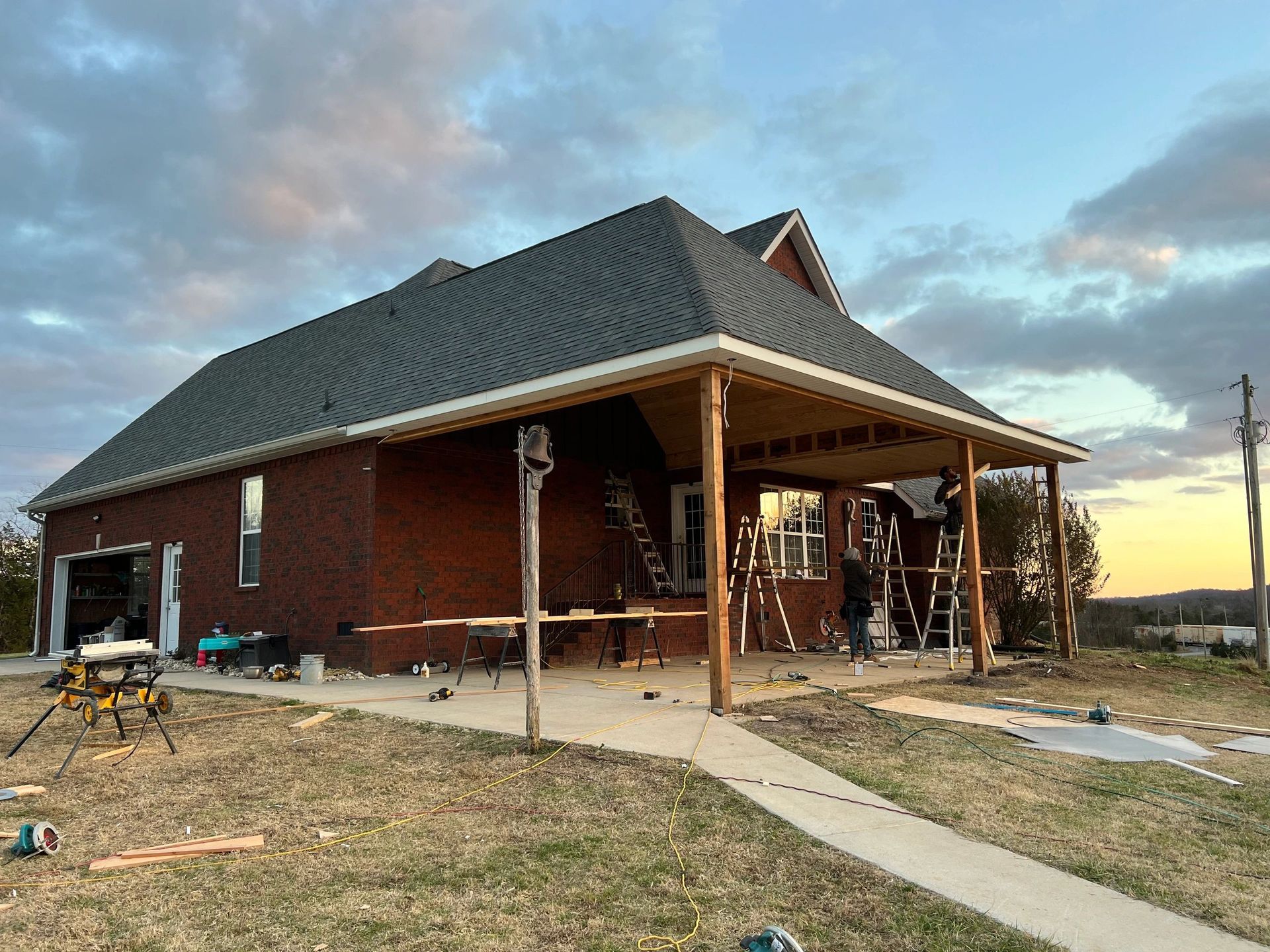 Home with porch under construction; brick, brown wood, and grey roof; cloudy sky.