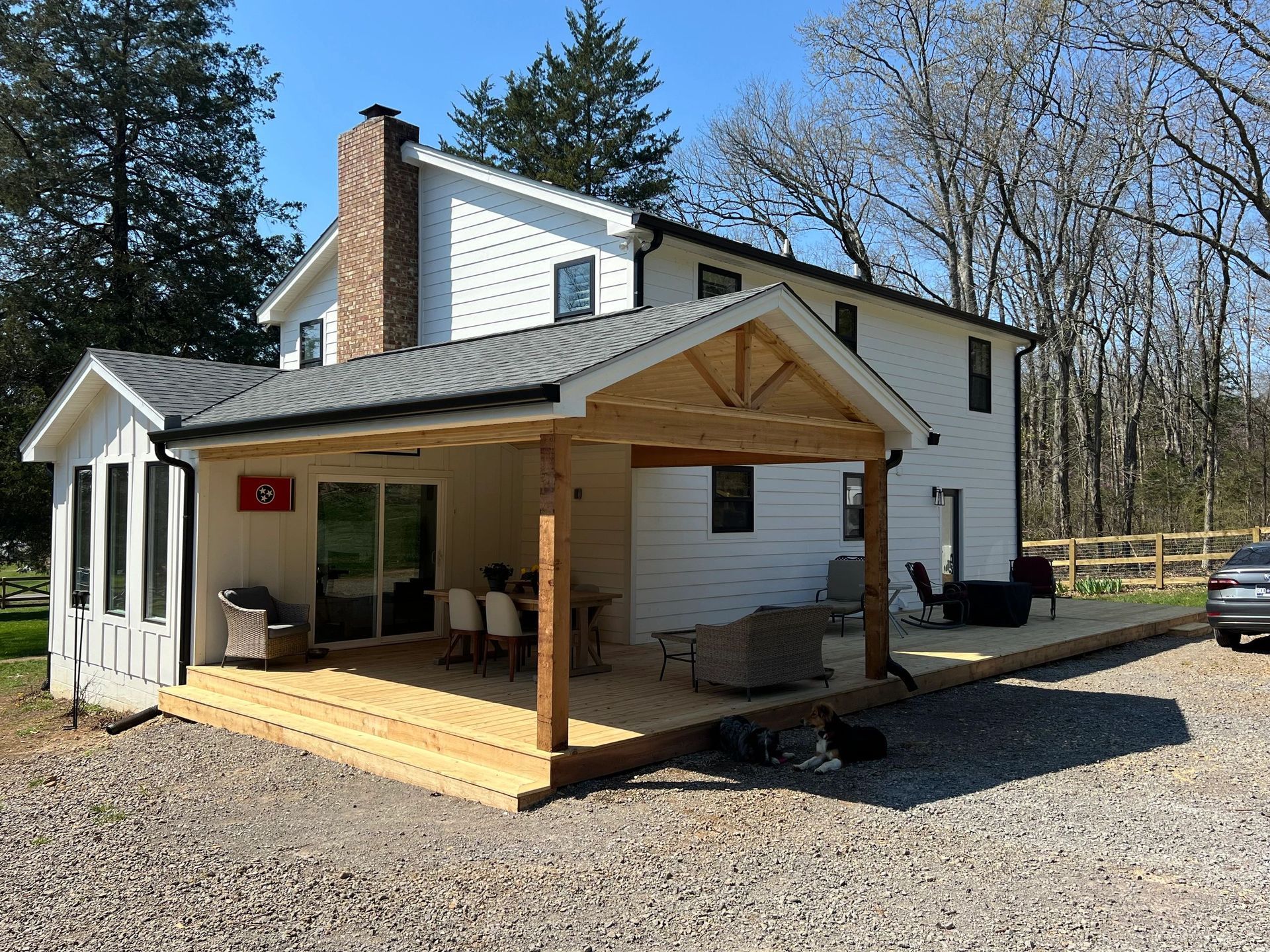 White two-story house with a wooden deck and porch. Trees in background, blue sky.