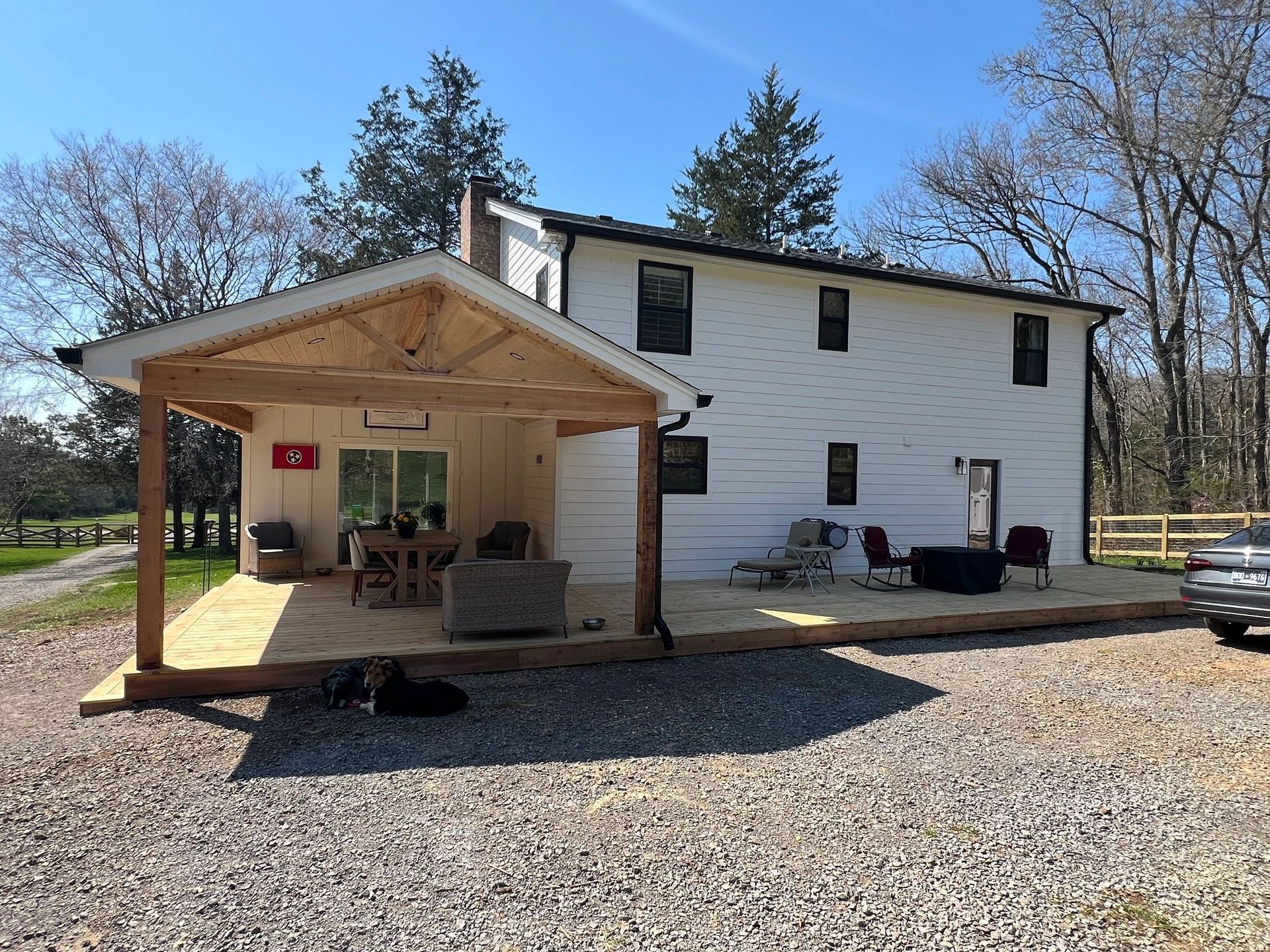 White house with a wooden deck, covered patio, and gravel driveway on a sunny day.
