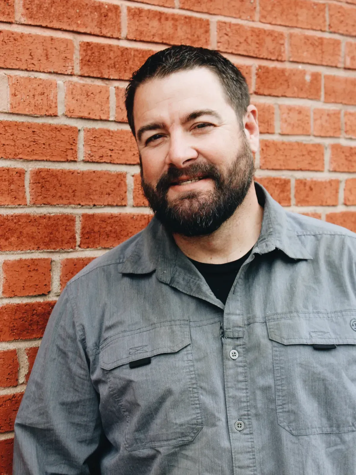 Man with beard leans against brick wall, wearing gray button-up shirt, smiles.