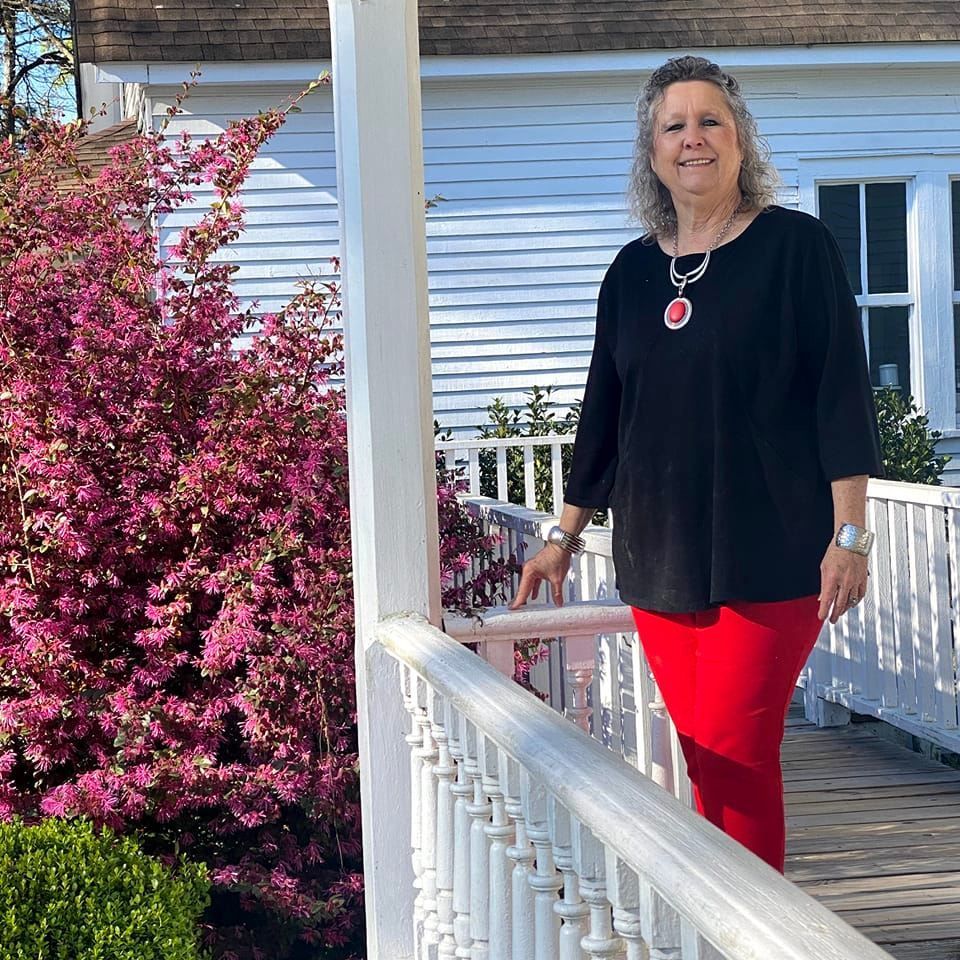Woman in red pants and black top on a porch with a flowering bush.