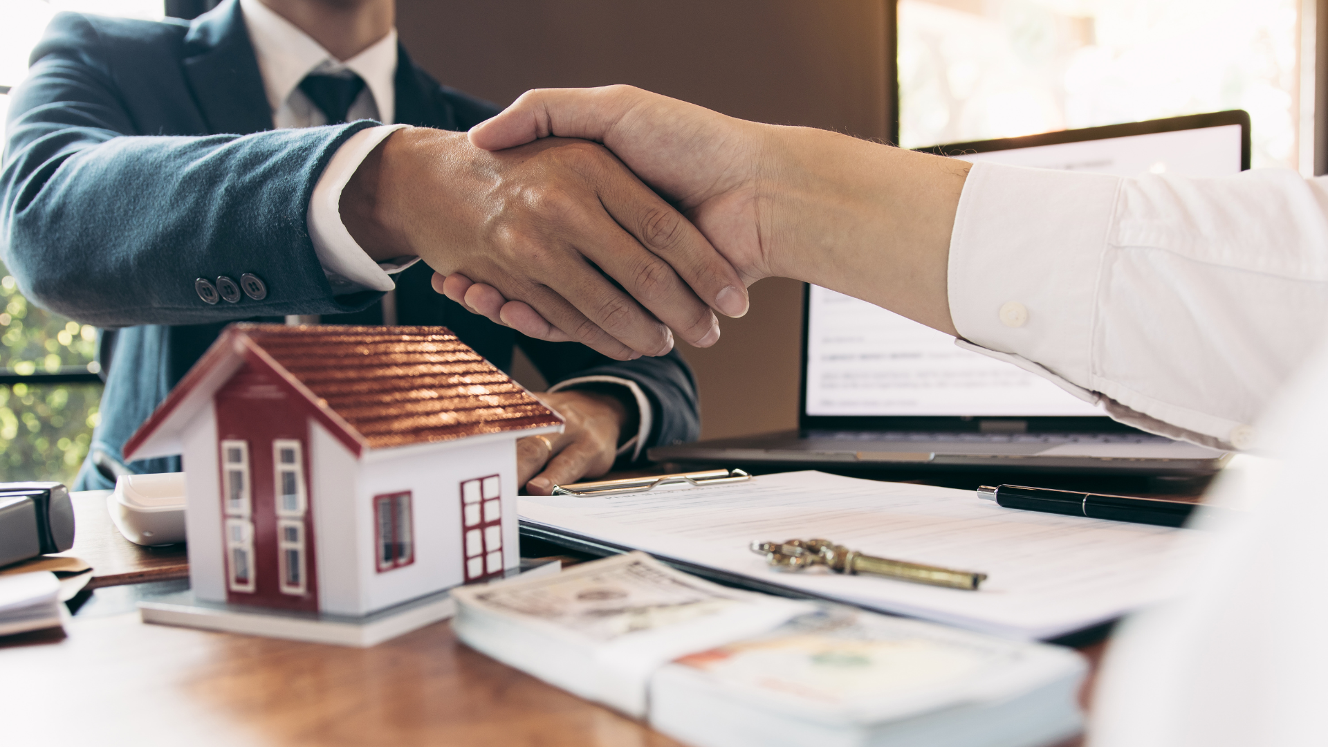 Two people shaking hands over a table with a model house, money, and paperwork.