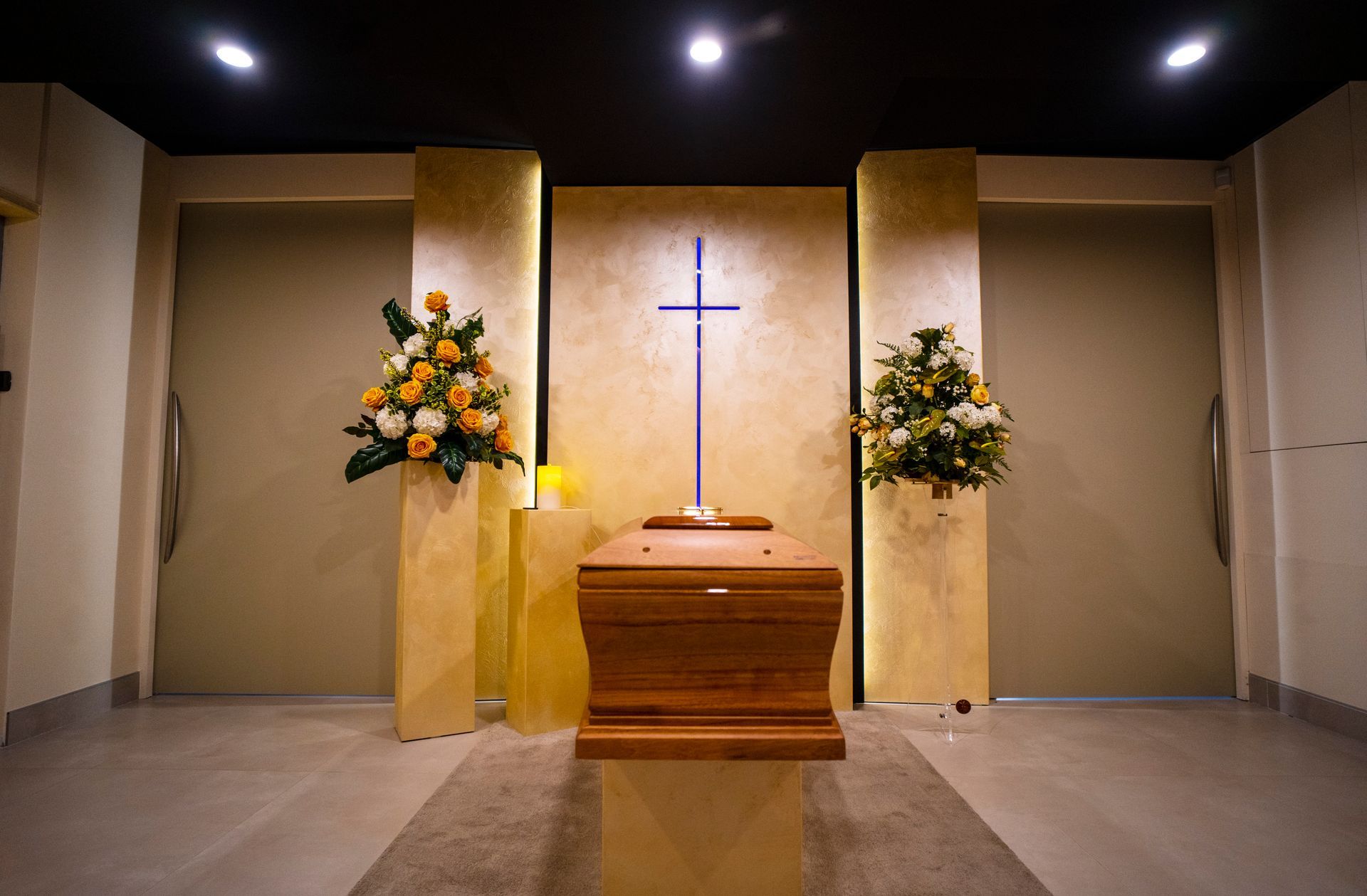 Una bara di legno è posta in una stanza con dei fiori e una croce.