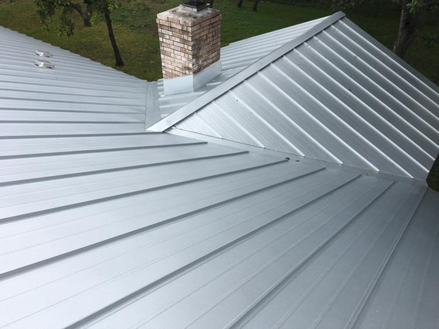 Silver metal roof with parallel ridges, brick chimney, and green landscape.