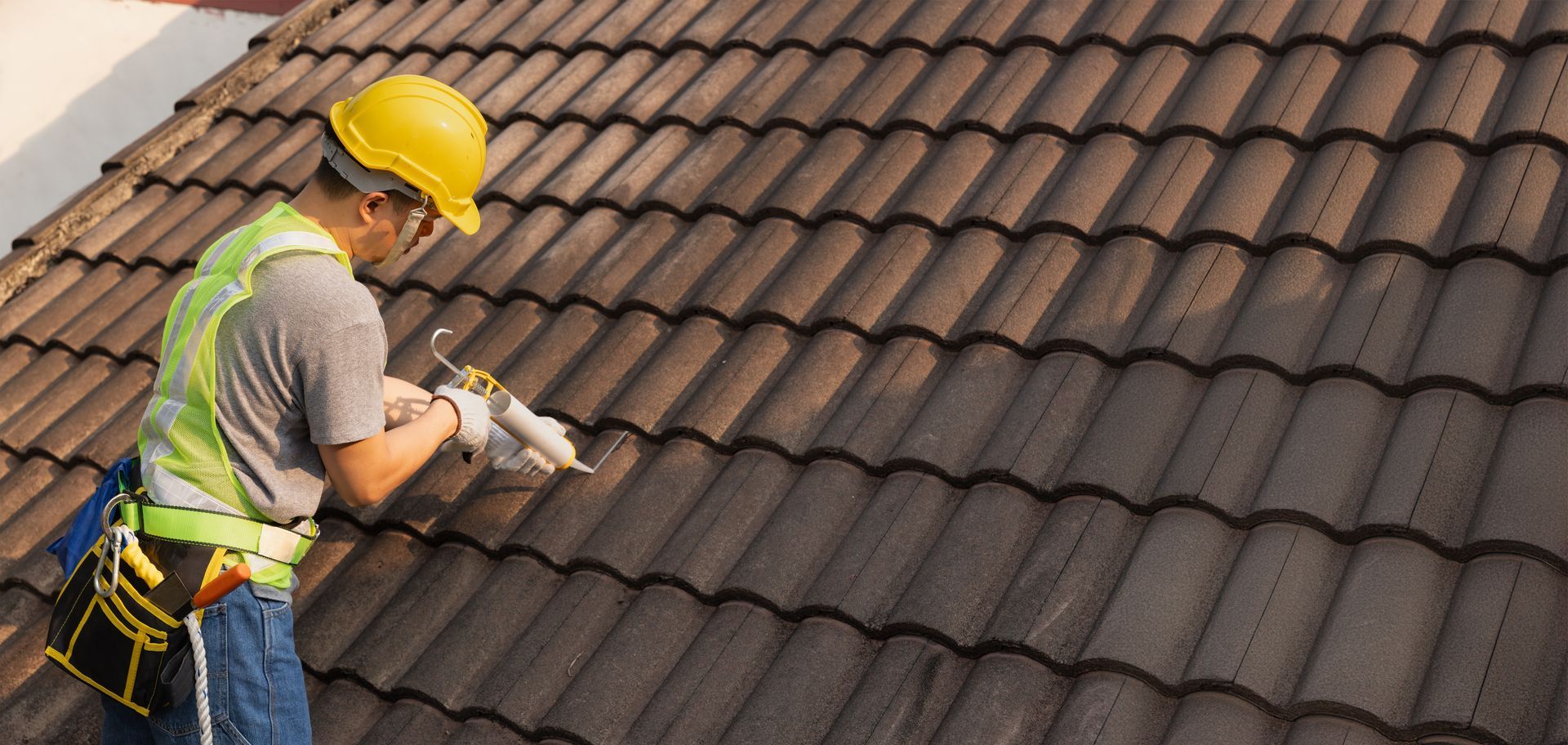 Roofer in yellow helmet and vest caulking roof tiles on a brown tiled roof.