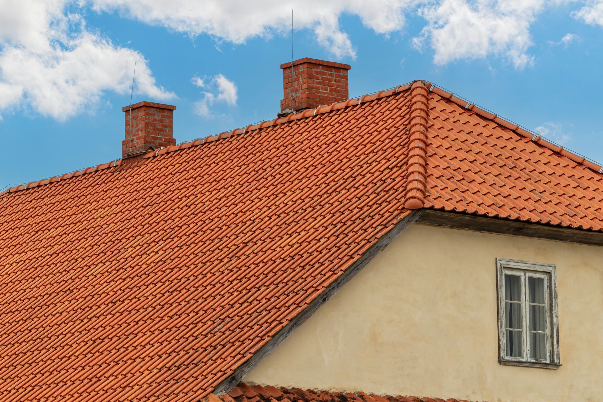 Red tile roof with two brick chimneys against a blue sky with white clouds. A tan wall with a window.