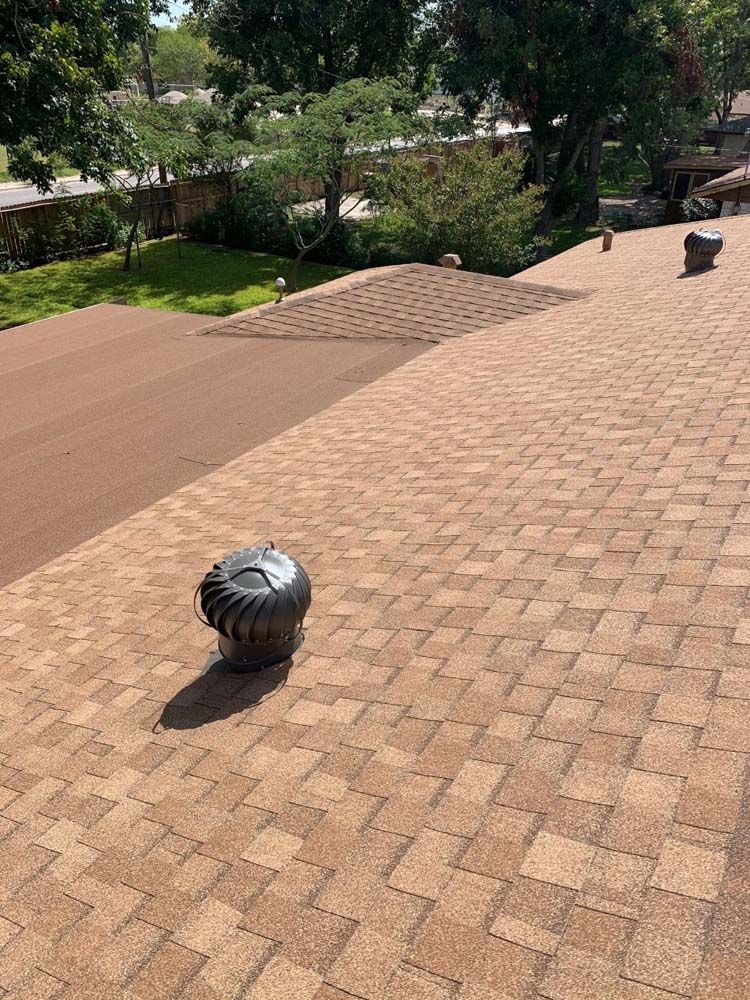 A brown shingle roof with a turbine vent, trees, and a small section of dark brown roofing.