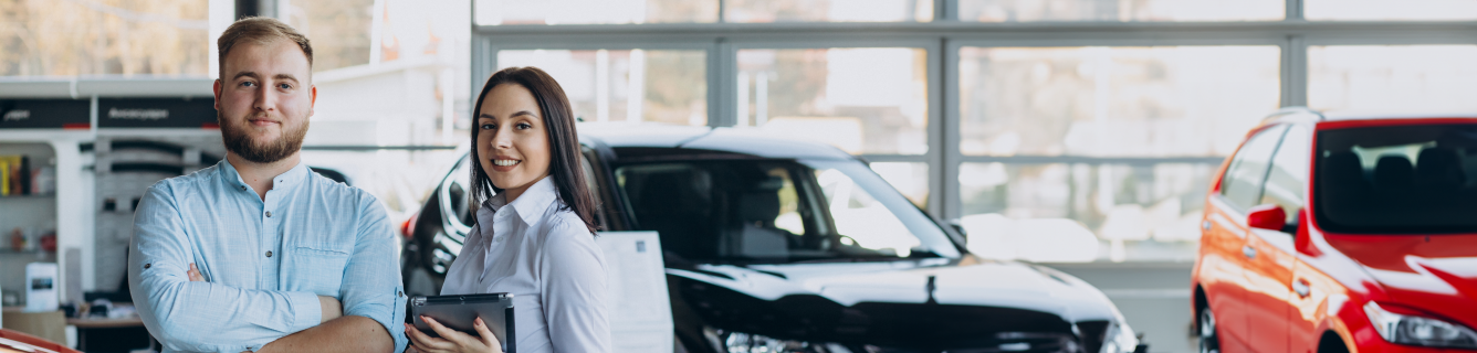 Dois vendedores de carros em uma concessionária. Um carro vermelho e preto é visível. A mulher segura um tablet.
