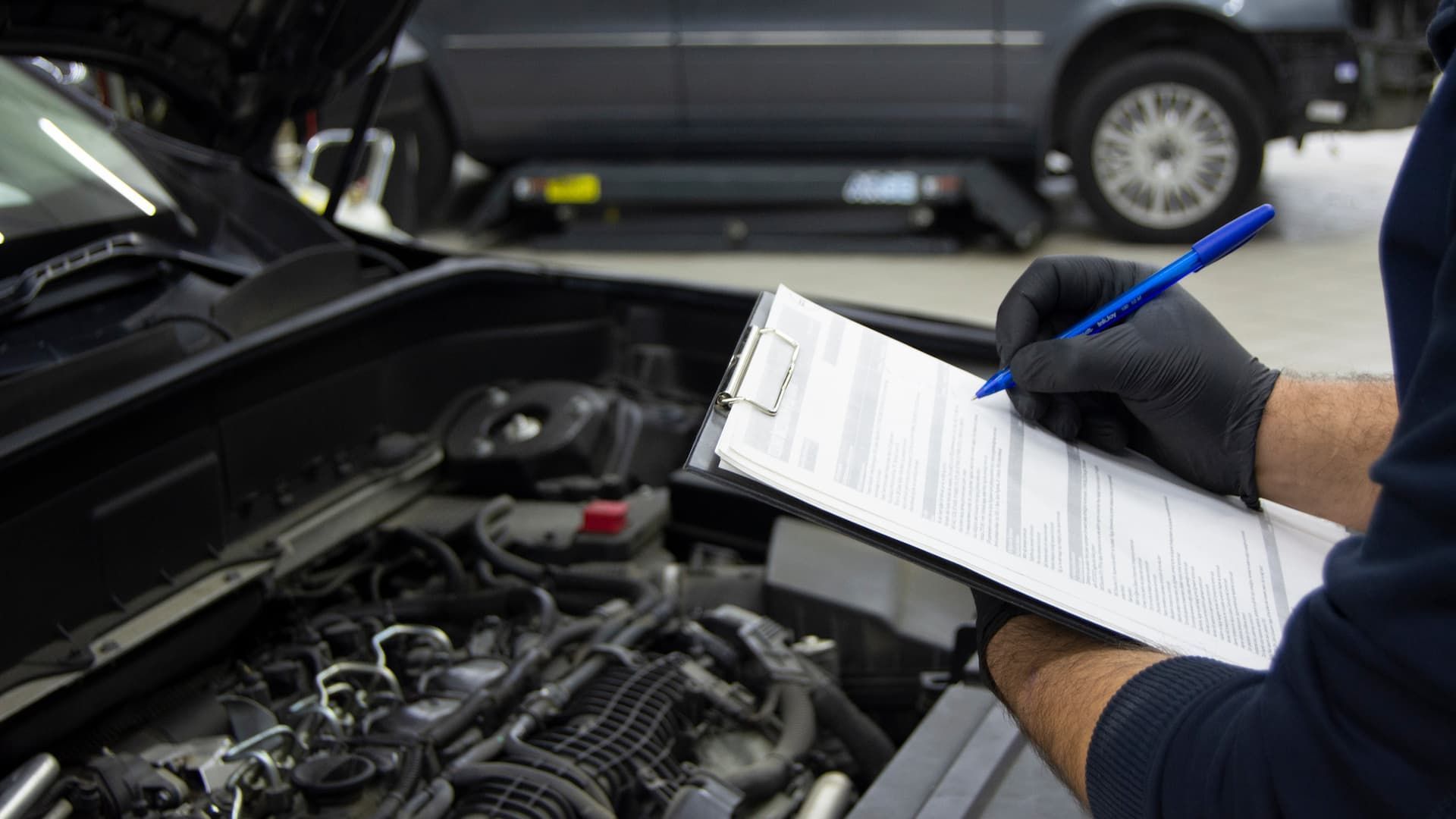 Mechanic writing on a clipboard beside an open car engine in a garage