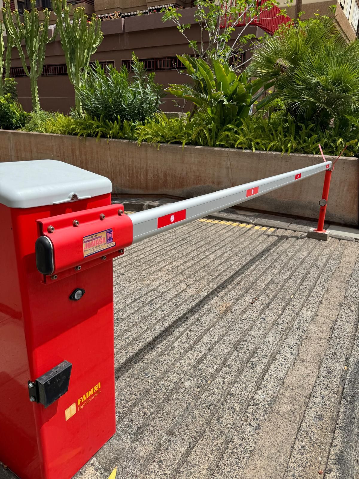 Barrera de aparcamiento roja con brazo elevado sobre una superficie de hormigón. Plantas al fondo.