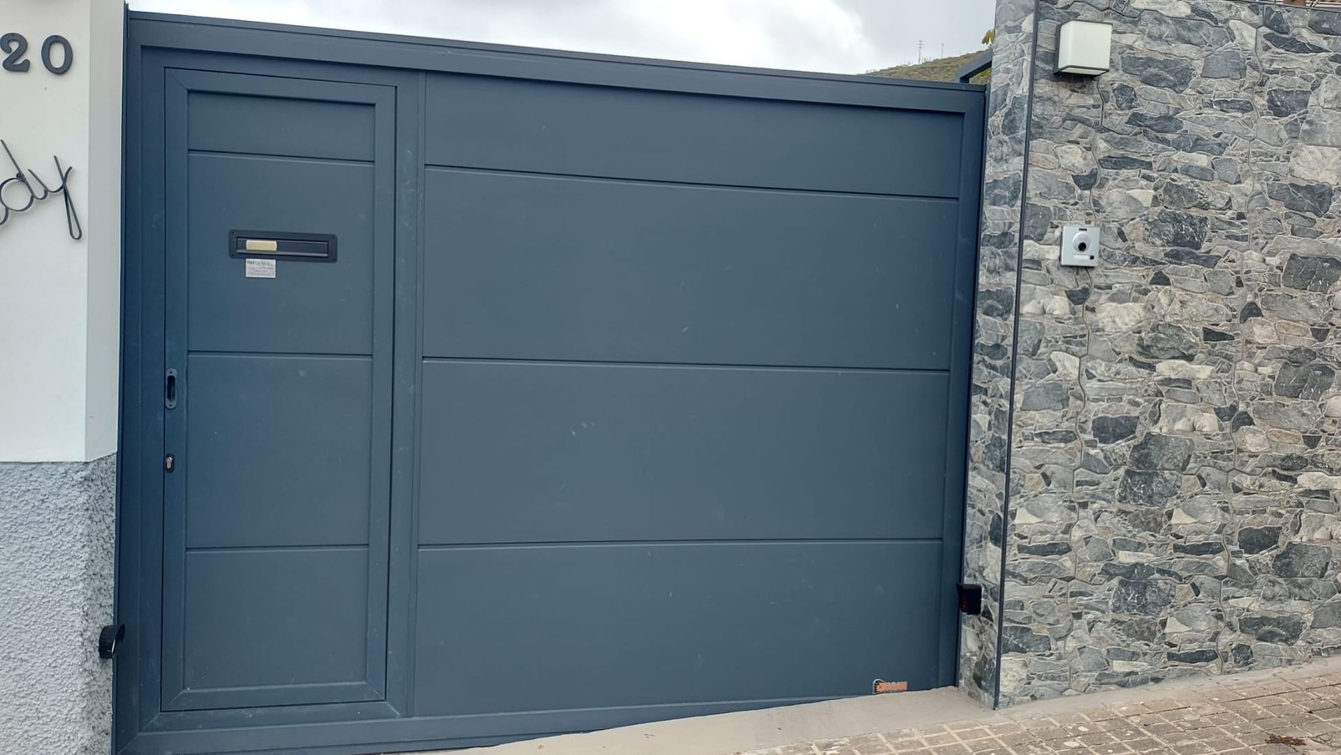 Portón metálico de color gris con pequeña puerta de acceso y buzón, empotrada en muro de piedra.