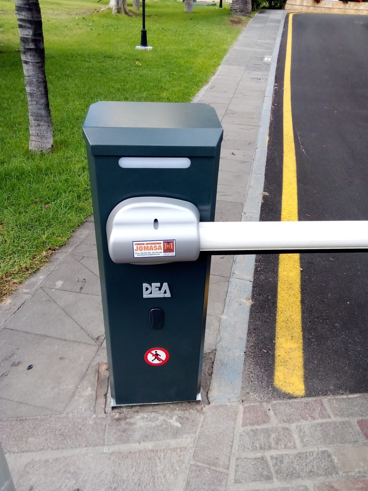 Barrera de aparcamiento automatizada de color verde oscuro con brazo blanco, situada en una acera junto a una carretera marcada con líneas amarillas.