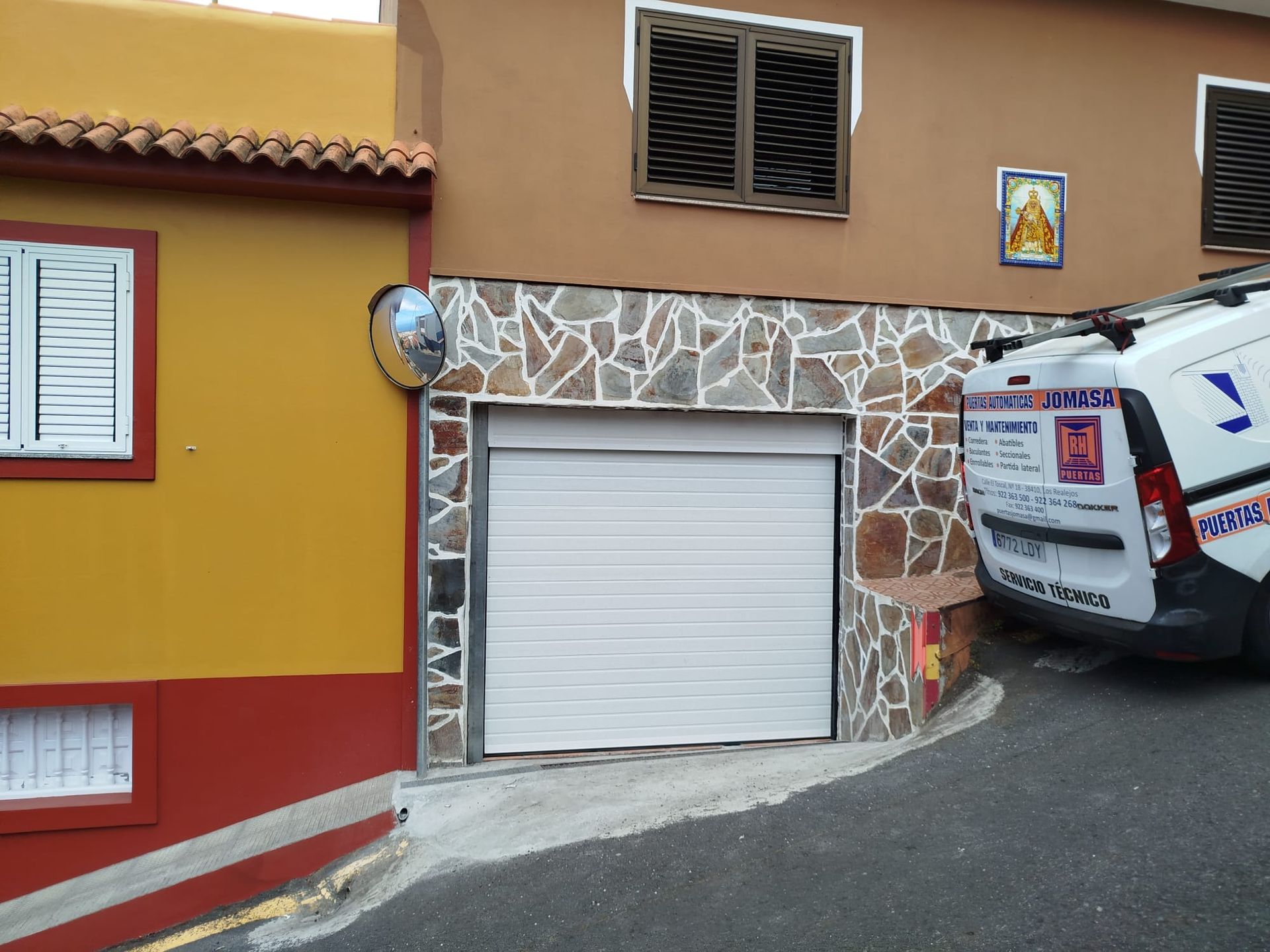 Puerta de garaje, blanca, debajo de edificio marrón con fachada de piedra; furgoneta estacionada en calle en pendiente.