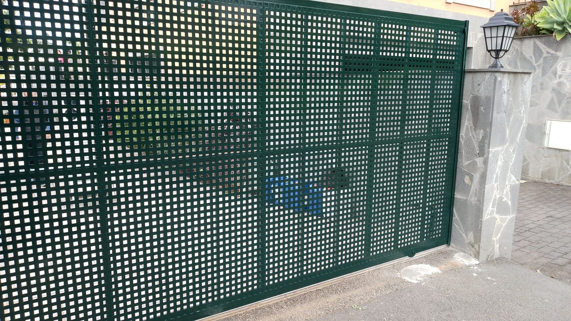Puerta de celosía verde sobre un poste de piedra con una lámpara, situada en un camino de entrada pavimentado.