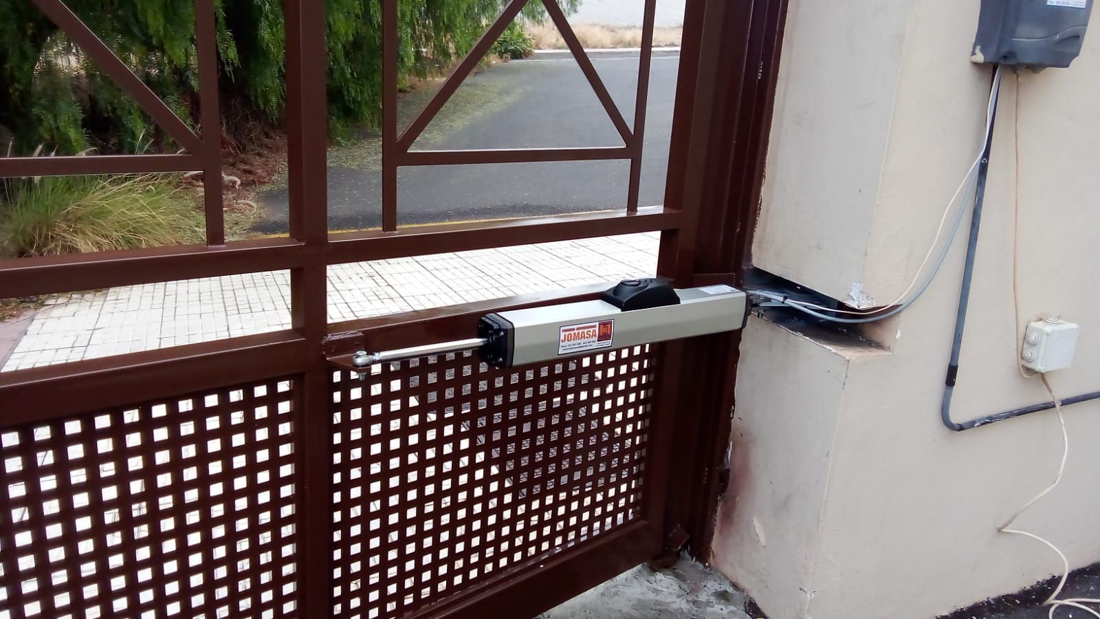 Puerta metálica marrón con abridor eléctrico adosada a una pared beige.