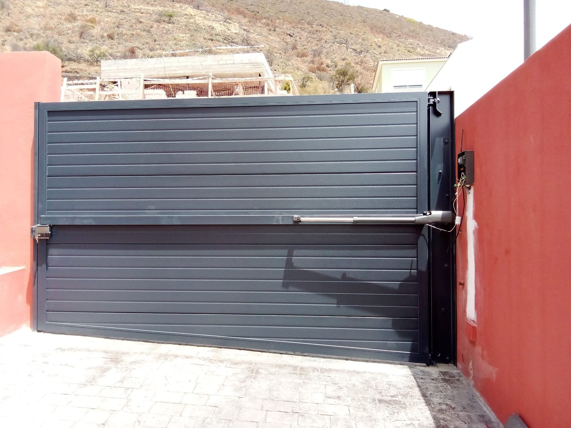 Portón metálico cerrado de color gris, con lamas horizontales, fijado a una pared de color rojo, con mecanismo en el lado derecho.
