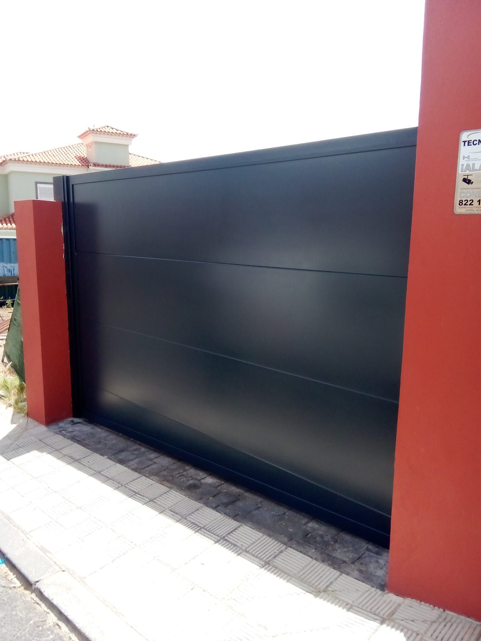 Puerta corrediza negra entre pilares rojos, frente a un edificio de color claro y una acera.