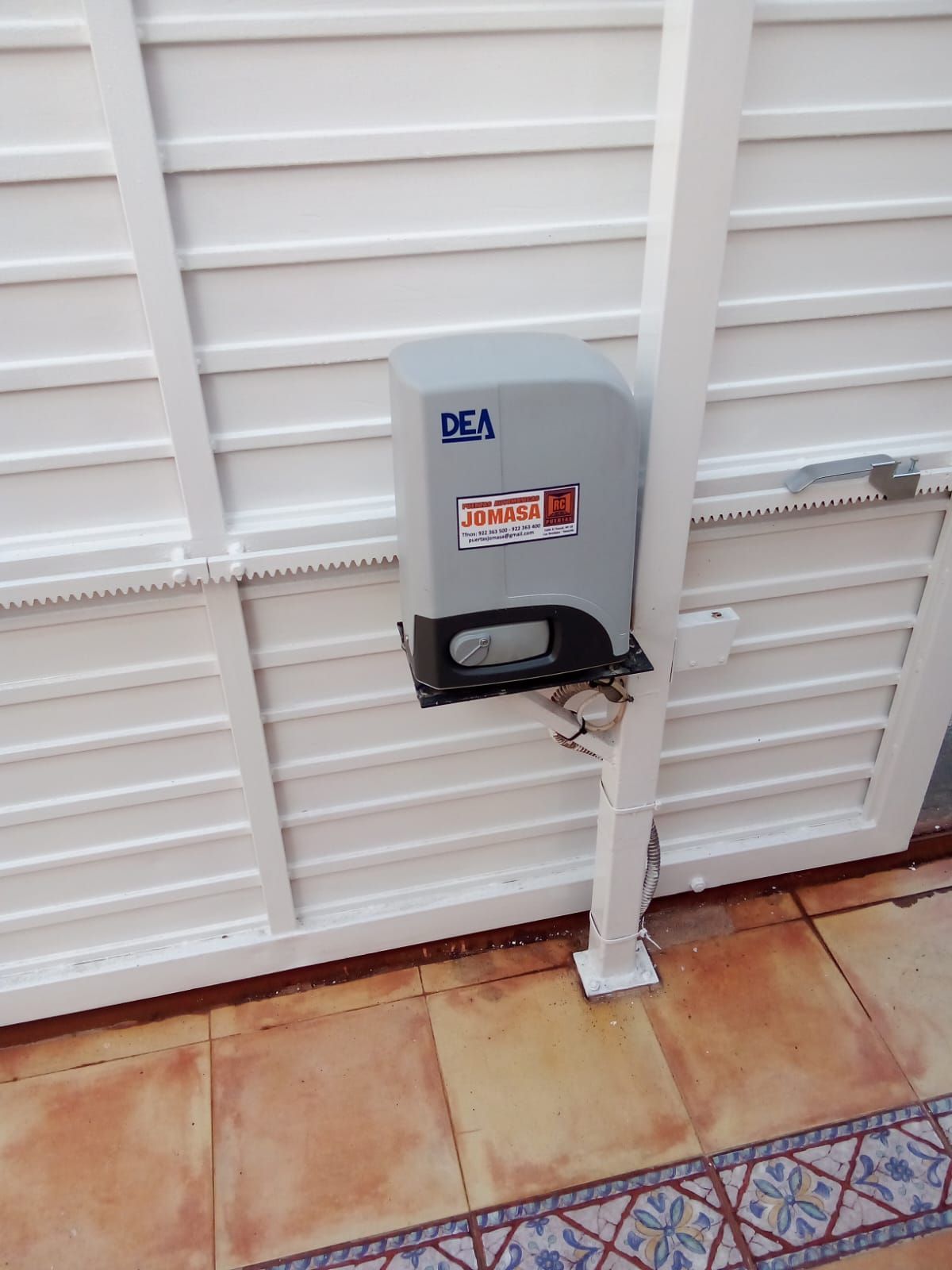 Abridor automático de puerta corredera montado sobre una puerta de metal blanca con suelo de baldosas de color beige.