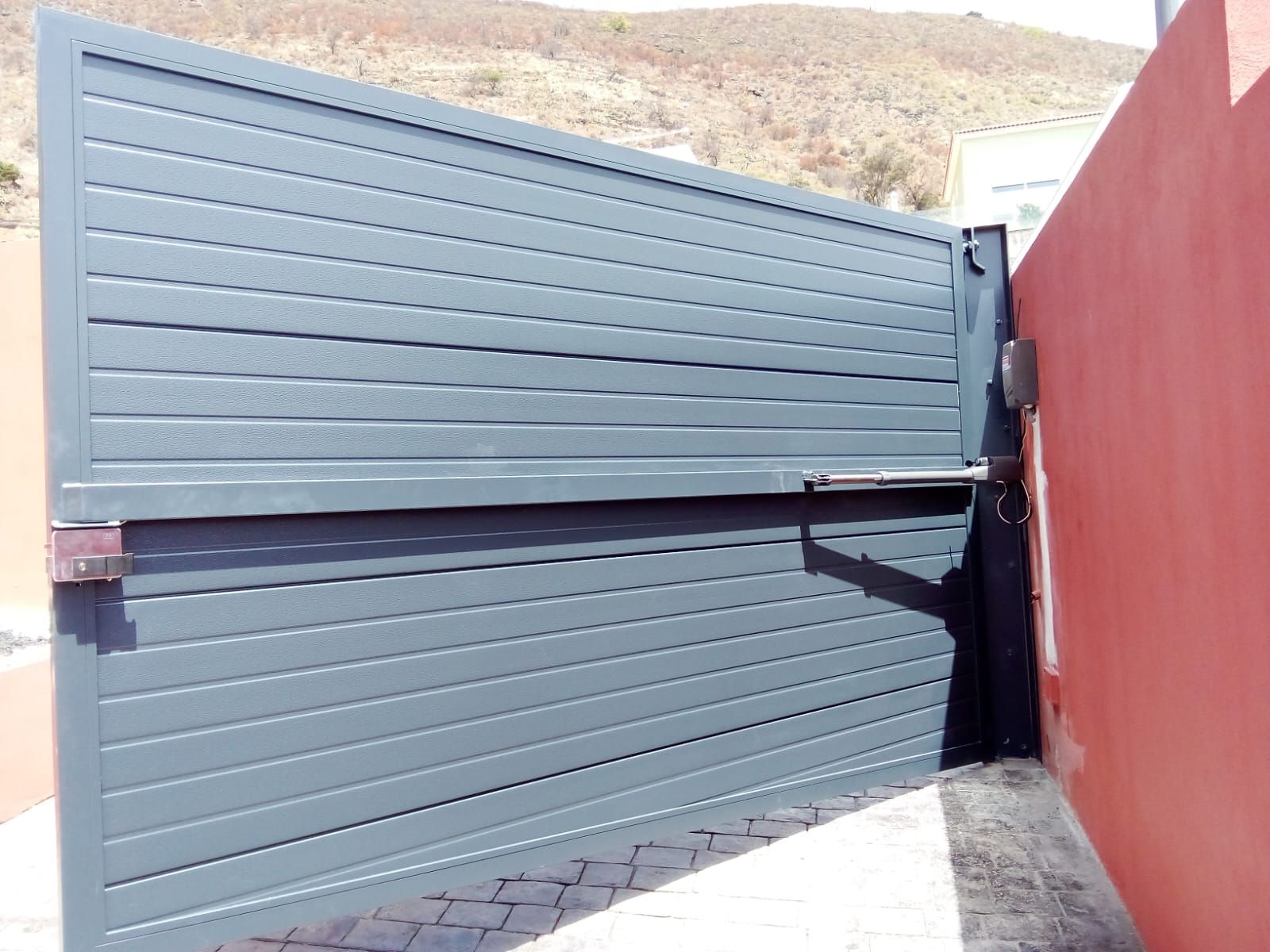 Puerta metálica gris que se abre, junto a una pared roja, en un entorno exterior soleado.