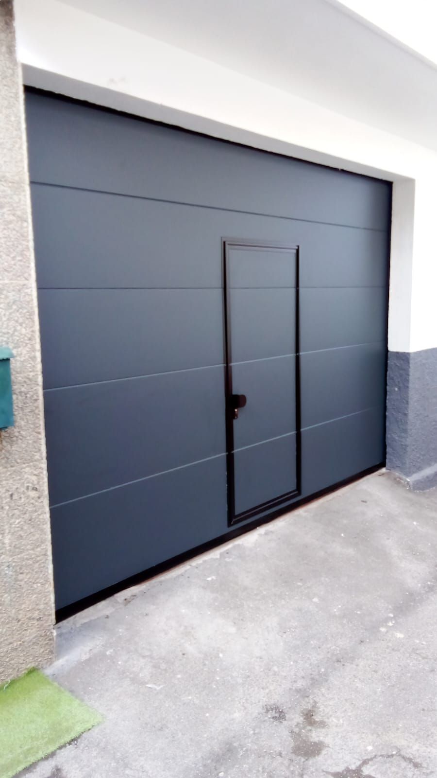 Puerta de garaje de color gris con puerta peatonal incorporada, empotrada en pared de hormigón.
