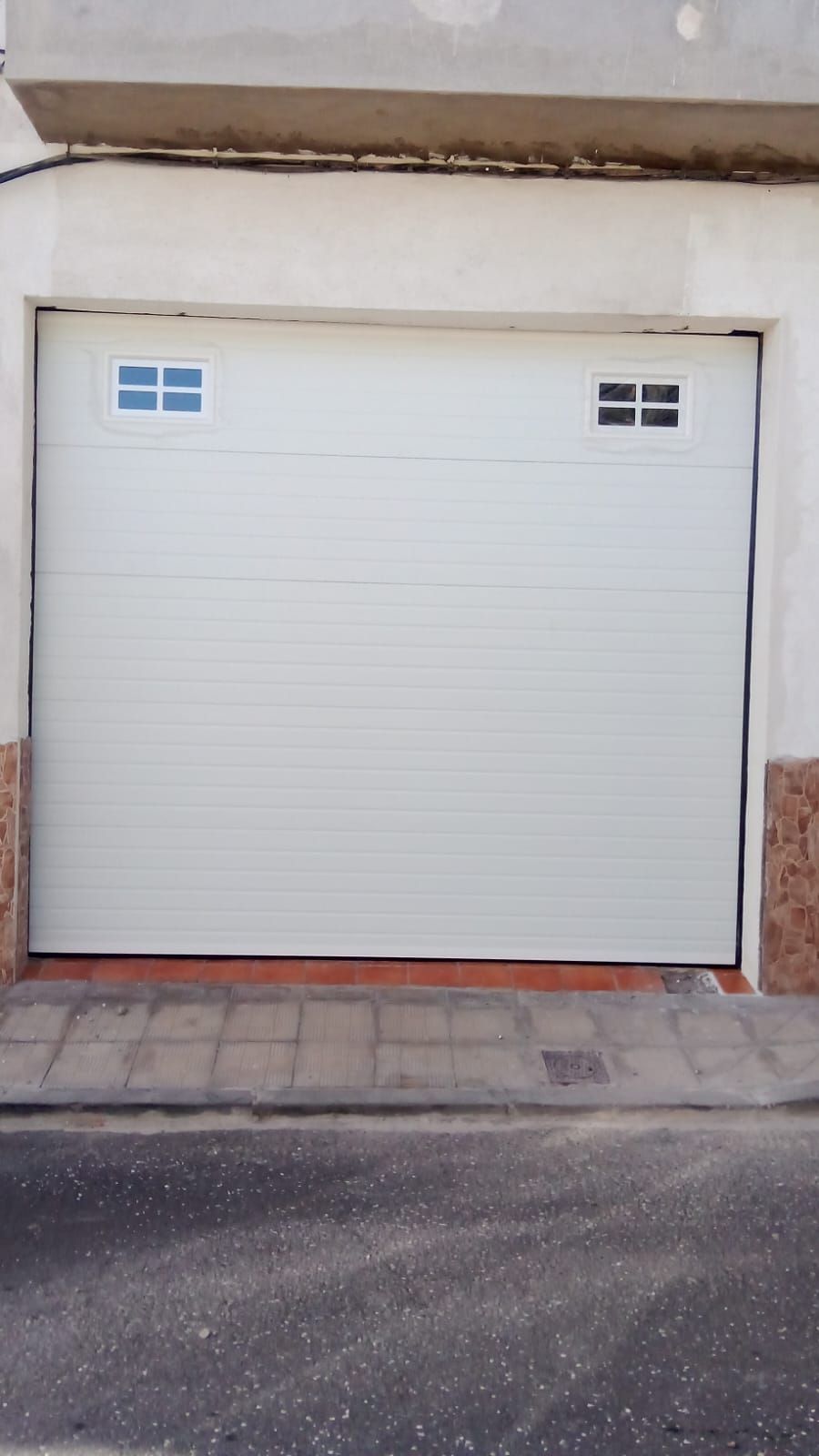 Puerta de garaje blanca con dos pequeñas ventanas cuadradas, ubicada en un edificio blanco con detalles de ladrillo.