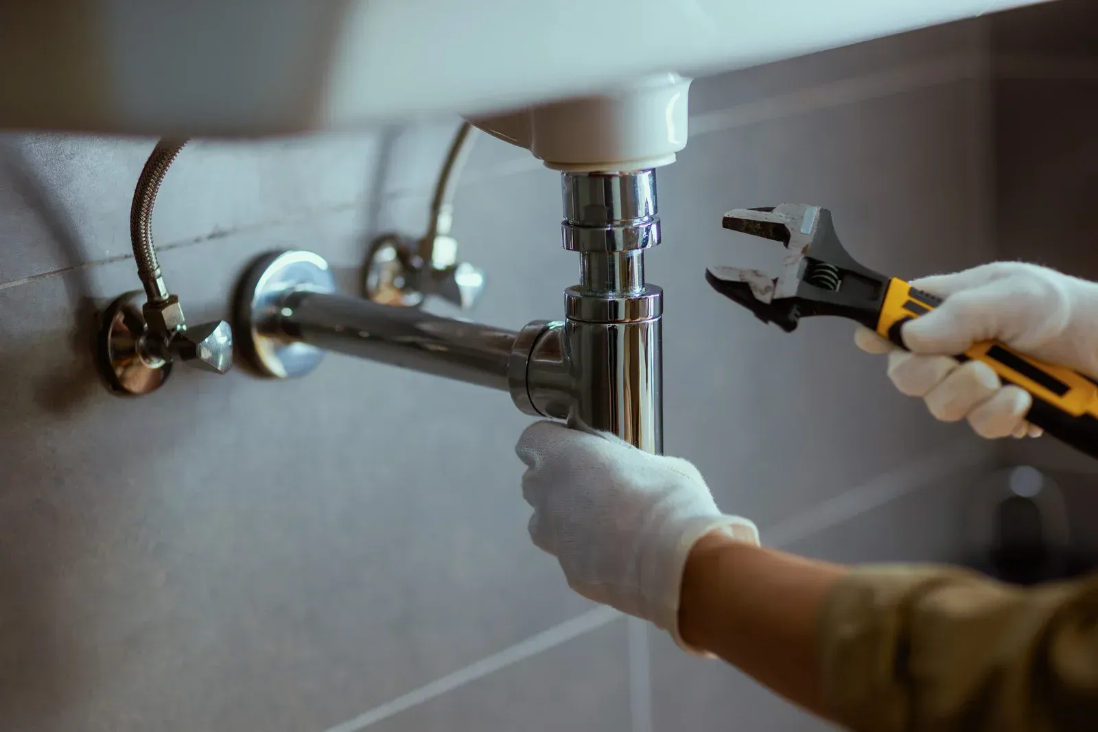 Plumber in white gloves using a wrench on a bathroom sink's drain pipes.